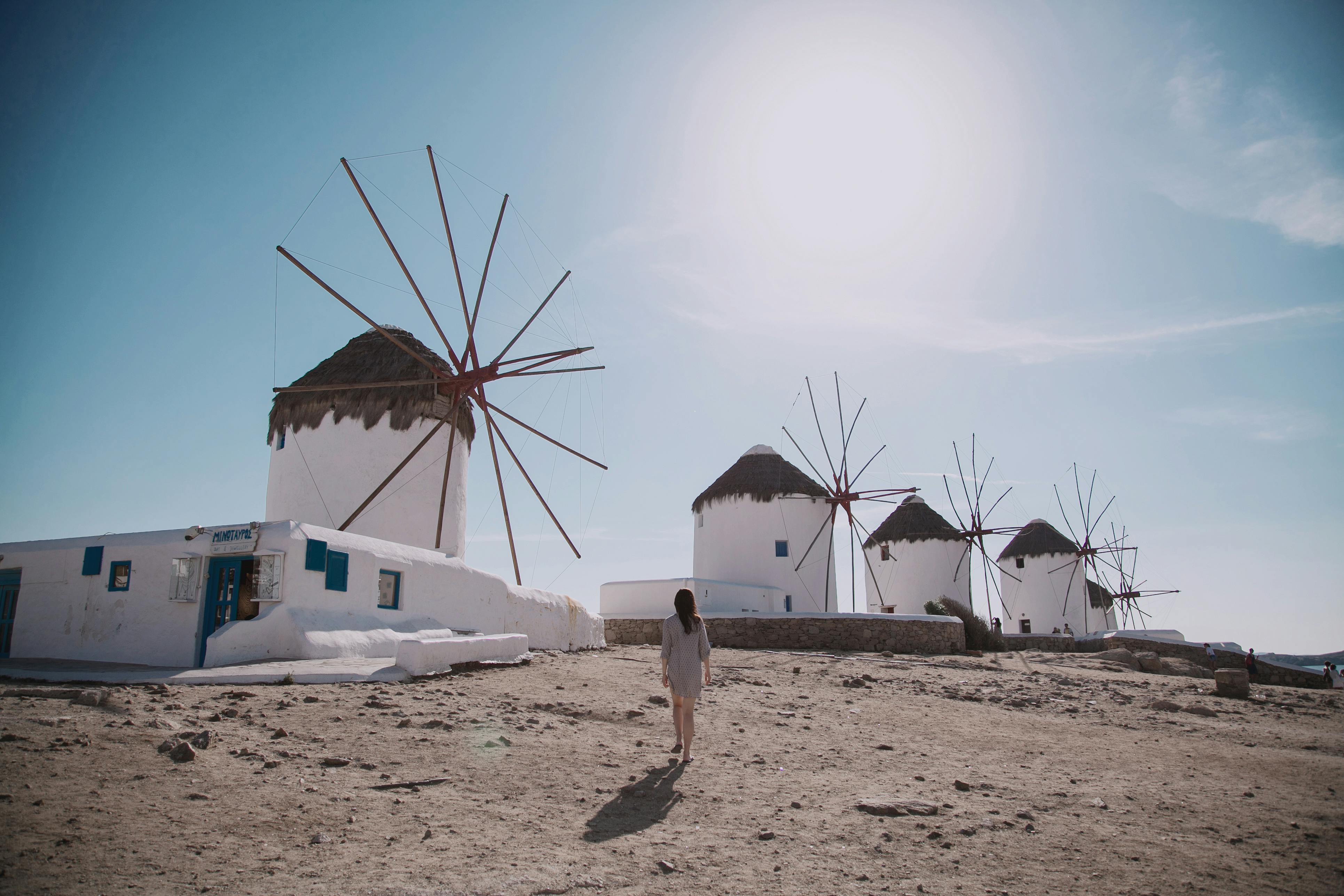 A woman walks toward the iconic white windmills of Mykonos under a bright sky, with sunlight casting long shadows on the ground.
