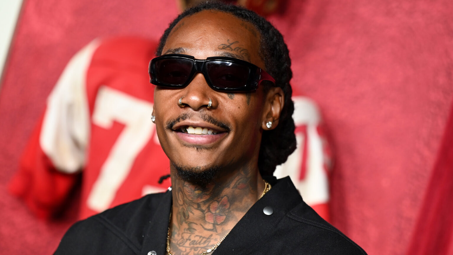 Wiz Khalifa with sunglasses, tattoos, and a nose ring smiles, wearing a black jacket. Red background with a blurred jersey.