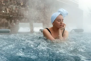 shutterstock_2437066017 Beautiful young woman relaxing in outdoor hot thermal pool. Snowy winter day. Thick steam. Enjoying Spa on holidays. High quality photo