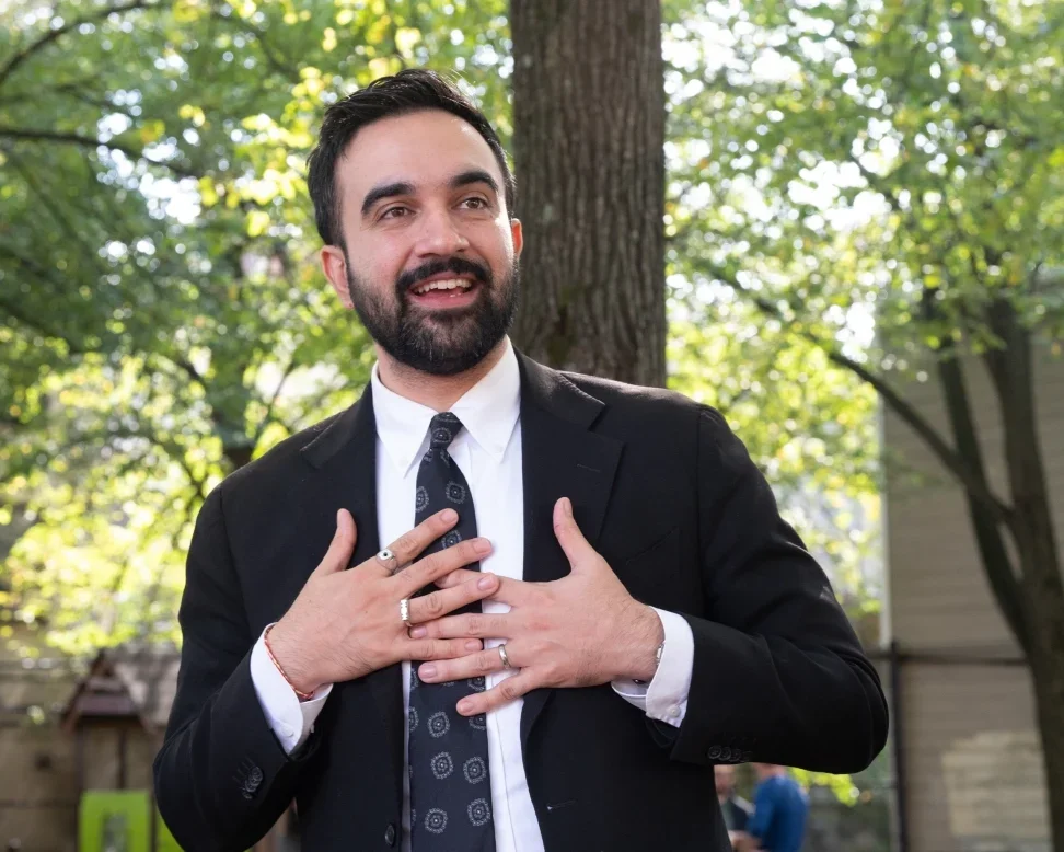 Zohran Mamdani smiling and looking off camera holding both hands to his chest. He is outside with trees behind him, and is wearing a suit and tie