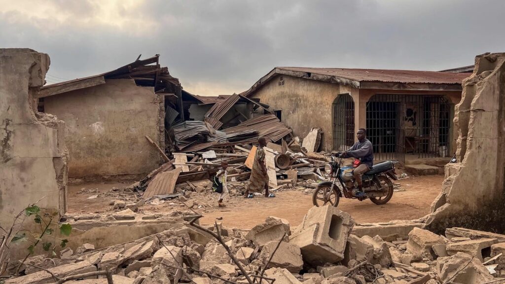 Residents and a motorcyclist move between destroyed structures in Offa on 27 December 2025 caused by debris from expended munitions that fell from US strikes on unspecified militants linked to the Islamic State group in Nigeria.