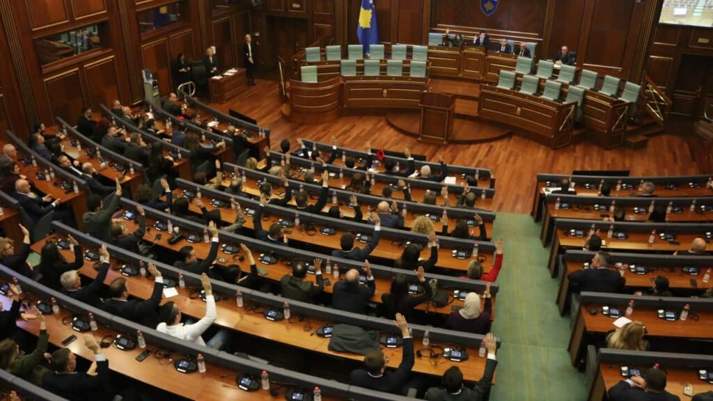 An elevated view of the Assembly of the Republic of Kosovo shows numerous members of parliament seated at curved wooden desks and raising their right hands to vote during a session.