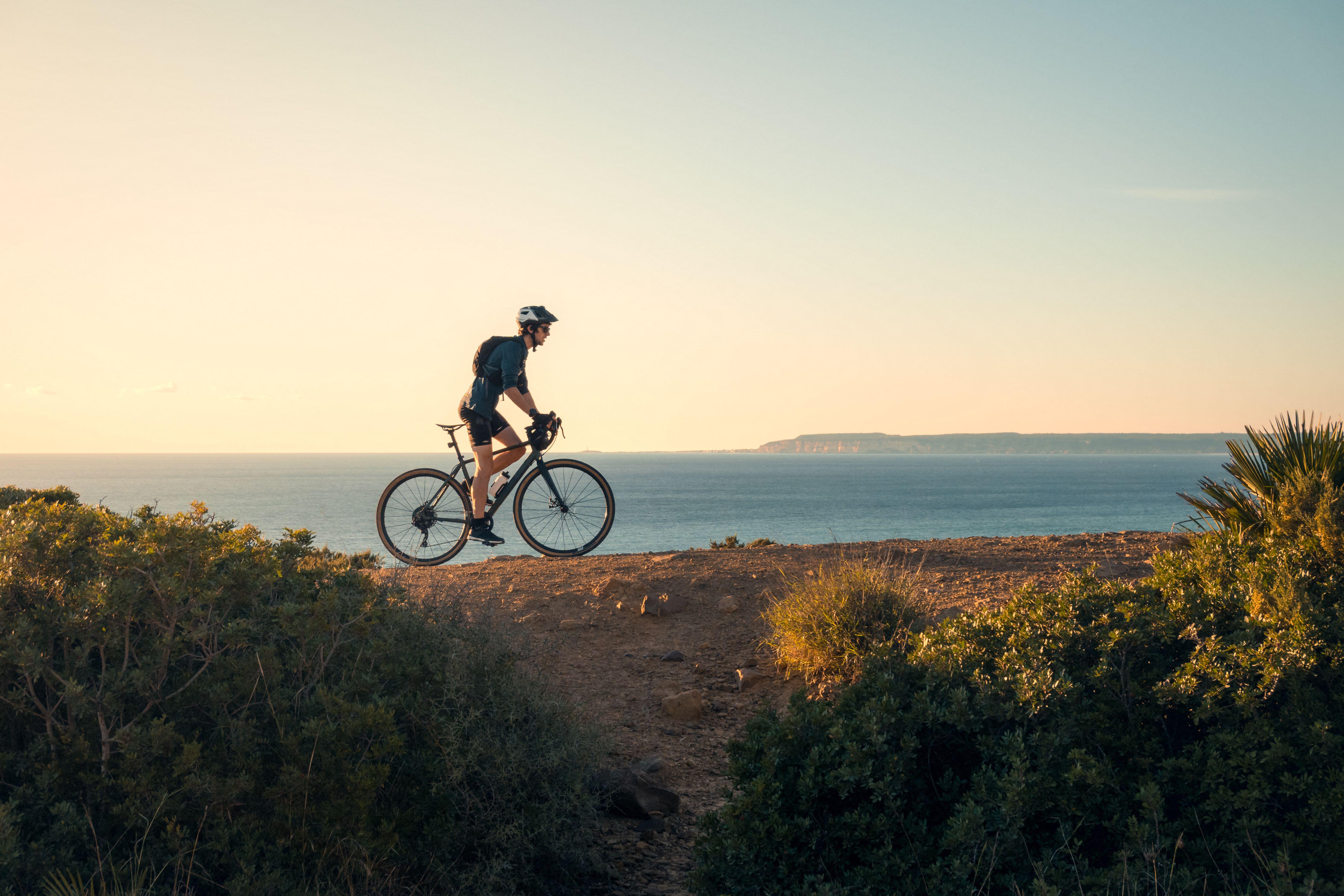 A young male cyclist rides along a scenic coastal path in Zahara de los Atunes, Cadiz, with the ocean and cliffs in the background.