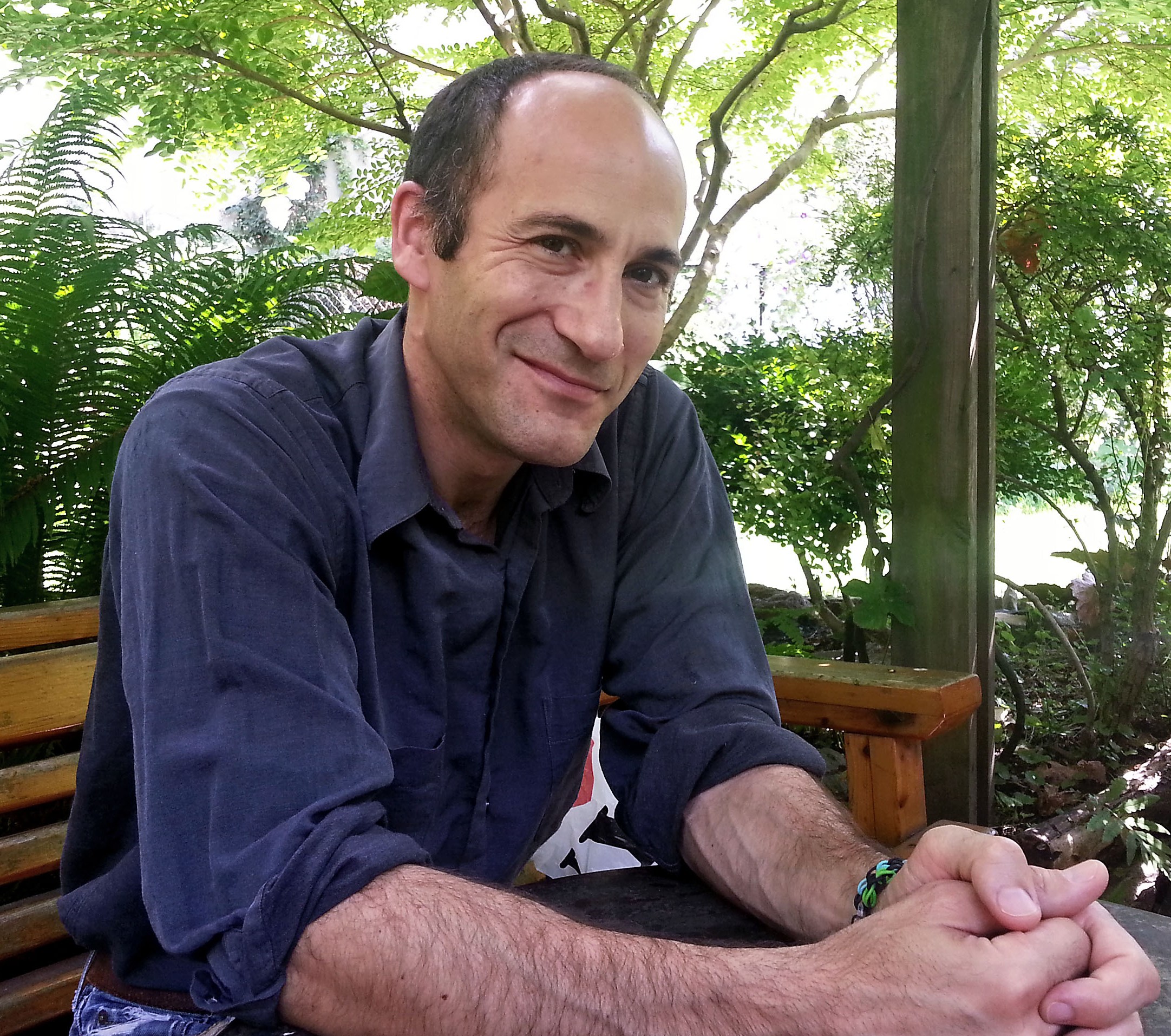 Adam Marcus smiling at the camera, seated at a wooden table outdoors with trees and ferns in the background.
