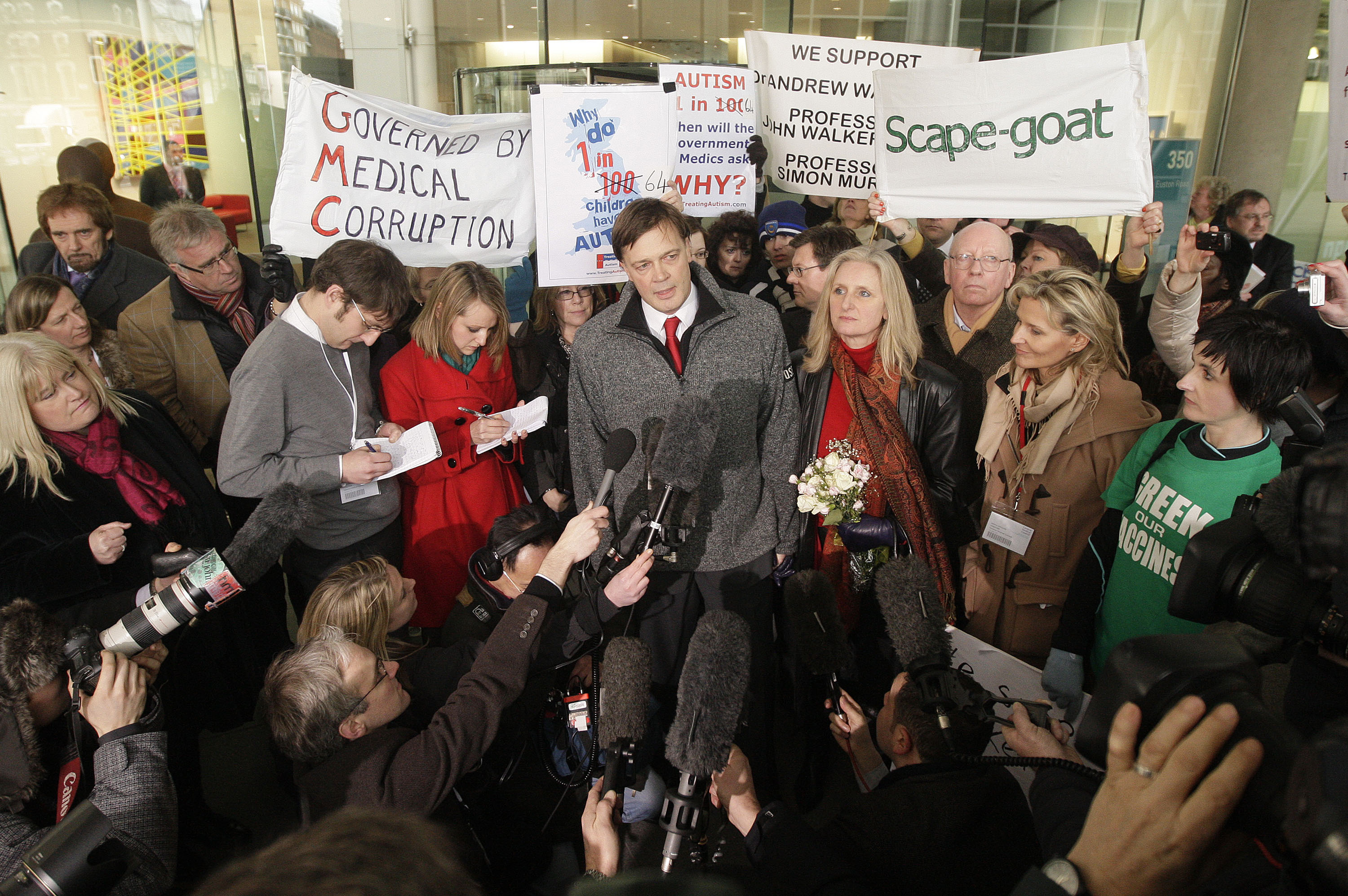 British doctor Andrew Wakefield speaks to media, surrounded by his wife Carmel, supporters holding signs, and reporters with microphones.