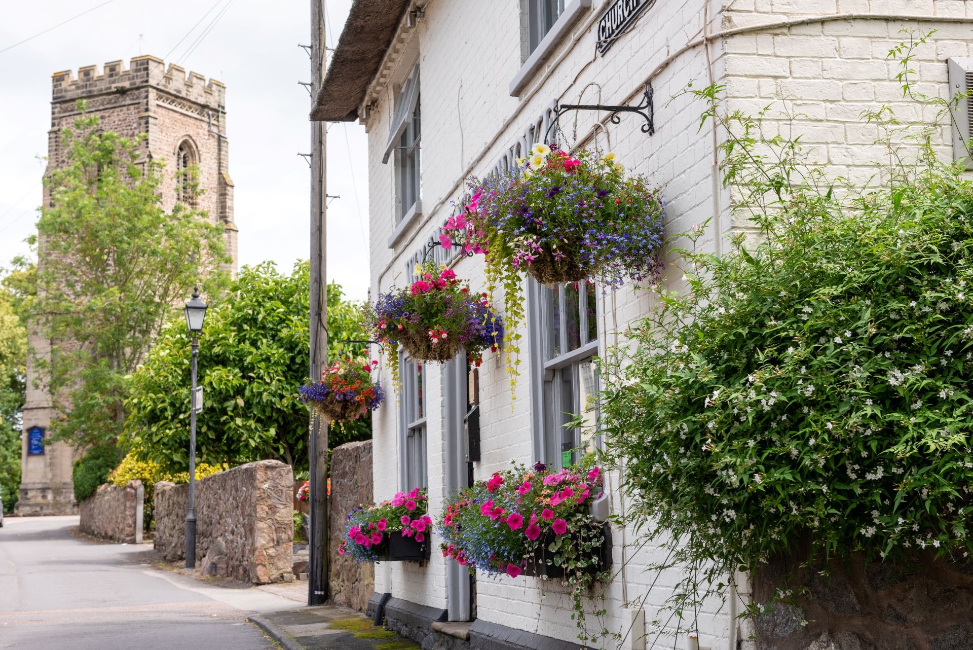 A white building with hanging flower baskets and window boxes, and a stone church tower in the background.