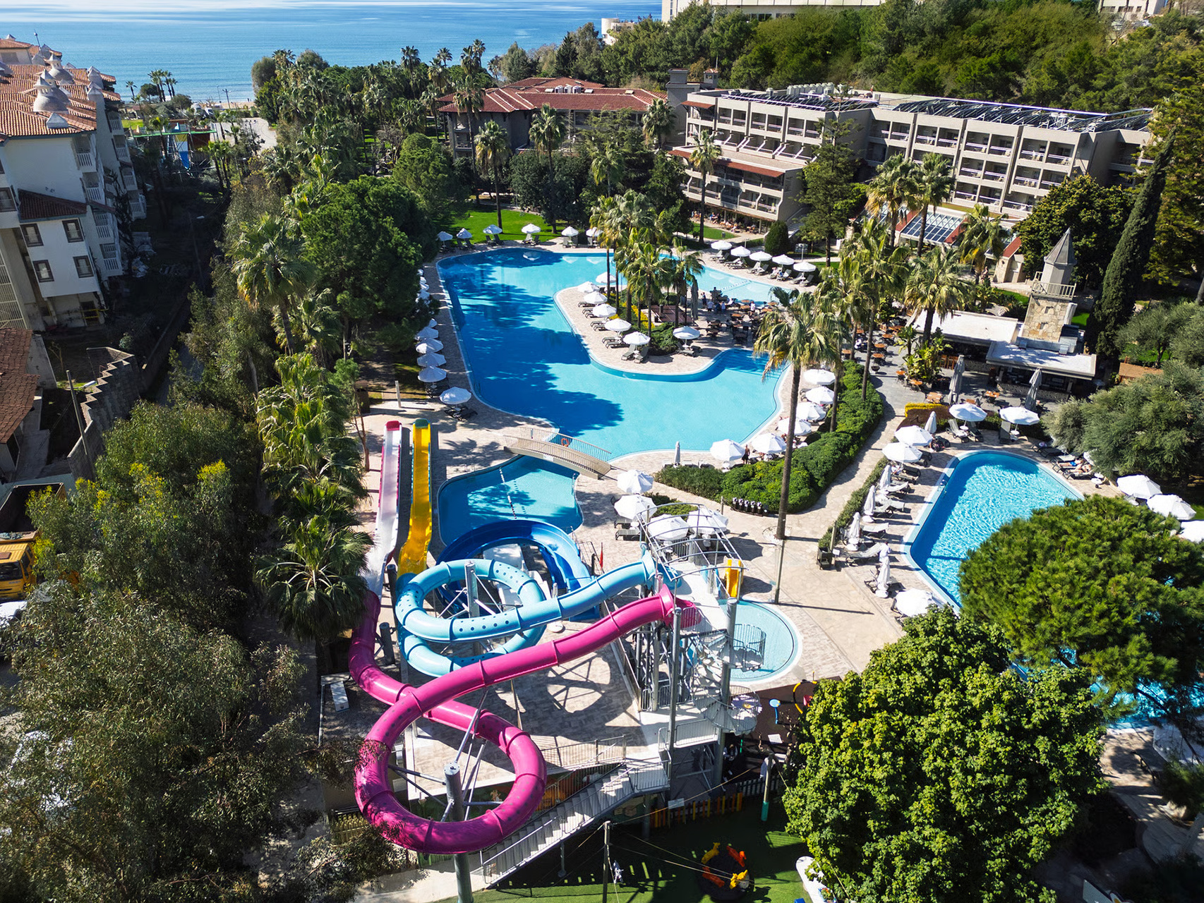 Aerial view of Barut Hemera, a resort with multiple pools, water slides, palm trees, and buildings, with the sea in the background.