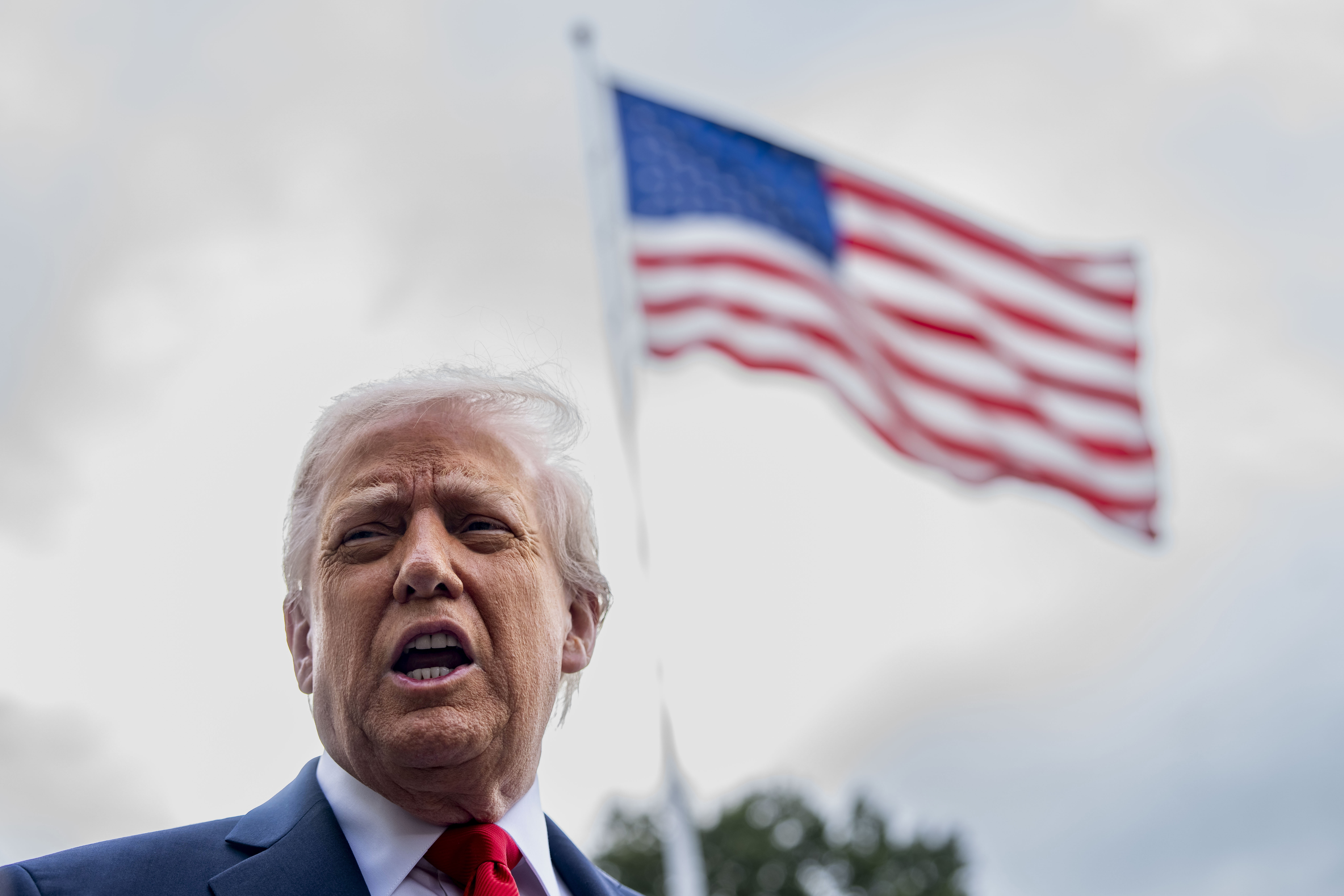 Donald Trump speaking to the press with an American flag in the background.