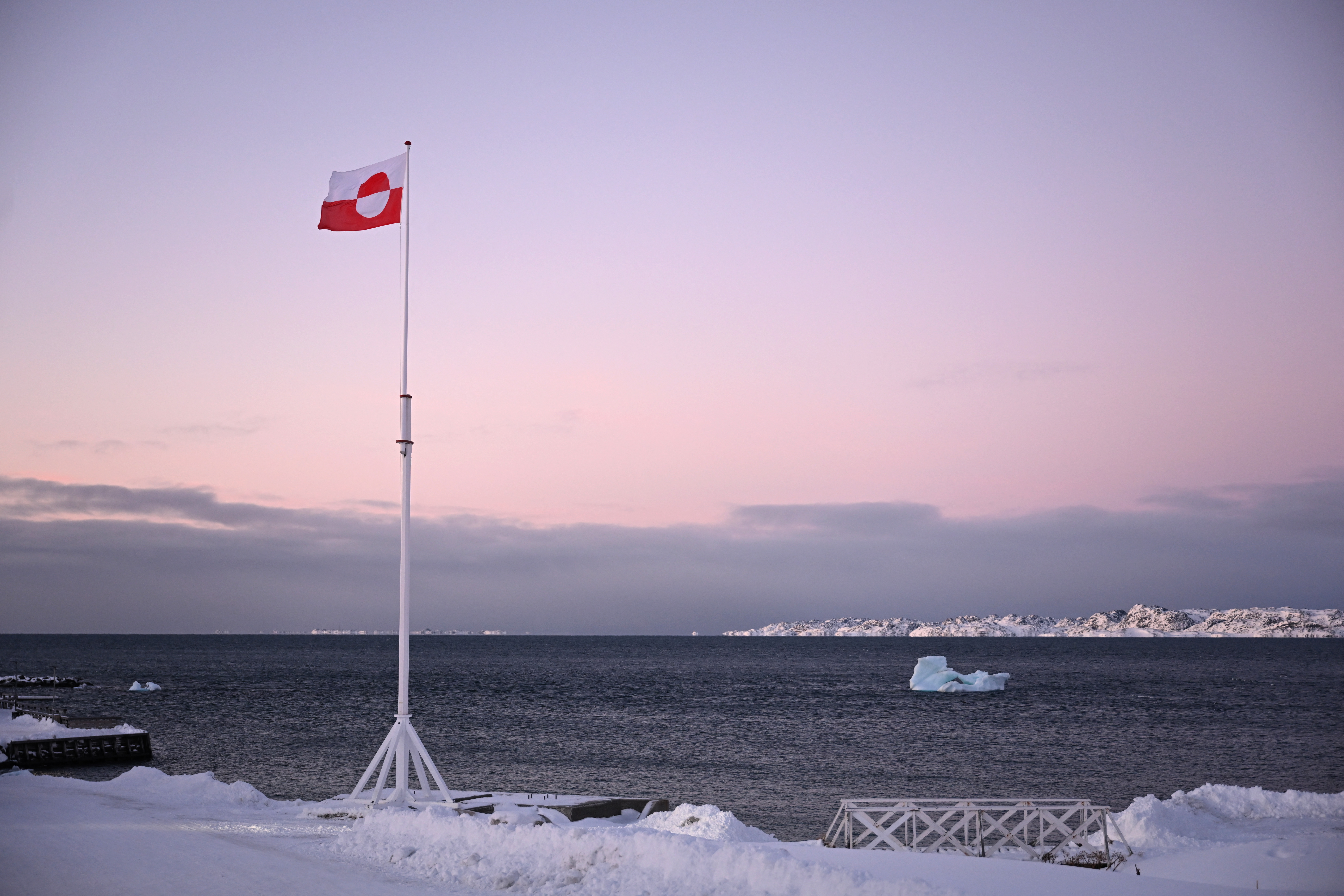 A Greenlandic flag flutters in the wind, with snowy ground and water with an iceberg in the foreground.