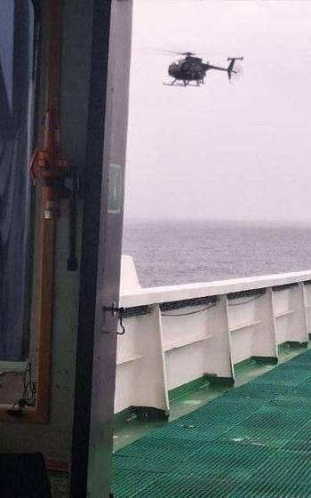 A military helicopter hovers over the ocean near a large ship, photographed from the deck which shows white railings and green textured flooring.