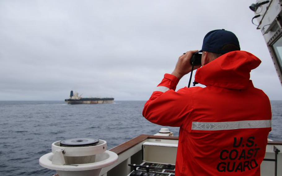 A U.S. Coast Guard member in a high visibility red orange jacket and dark cap uses binoculars to look at an oil tanker in the distance on the open ocean. 