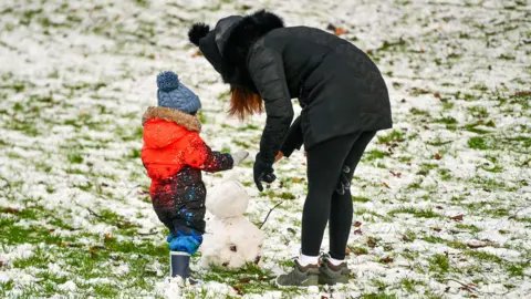 Pacemaker A woman and child in winter clothes stand around a small snowman. They are in a field covered in snow, with patches where the grass can be seen.