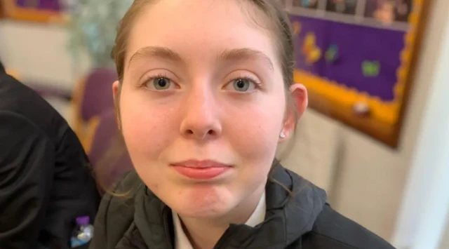 A young girl with a neutral expression on her face looks at the camera. She is in a school