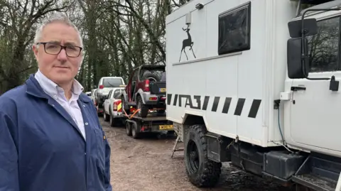 BBC A man with grey hair and black framed glasses is standing looking at the camera. He is wearing blue overalls. He is stood next to a fleet of 4x4 vehicles. 