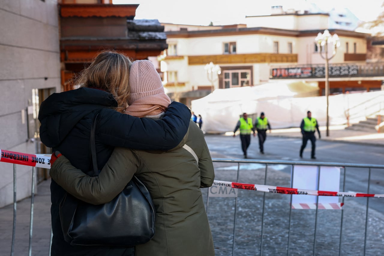 Two women in winter clothes embrace outside of a no-go zone cordoned off by police tape. Officers seen in the background. 