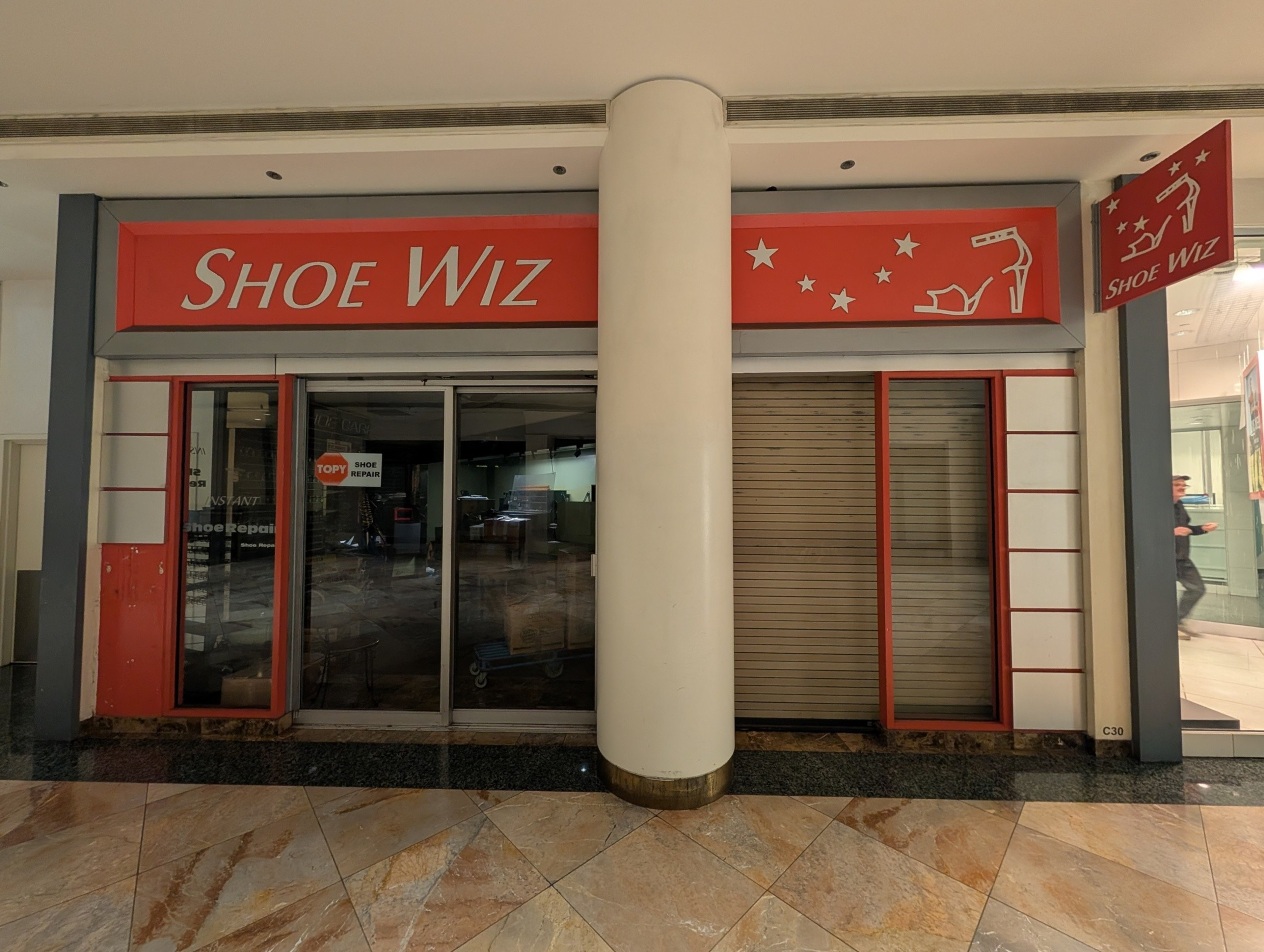 A closed shoe repair shop named “Shoe Wiz” has red signage with white text and shoe icons, glass doors, a metal shutter, and a large white pillar in front.