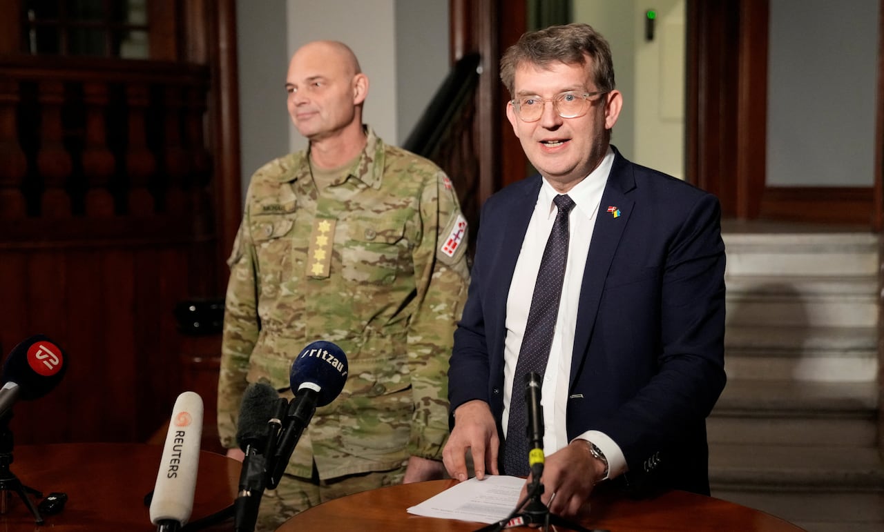 A man in a suit and another dressed in military fatigues stand at a news conference.