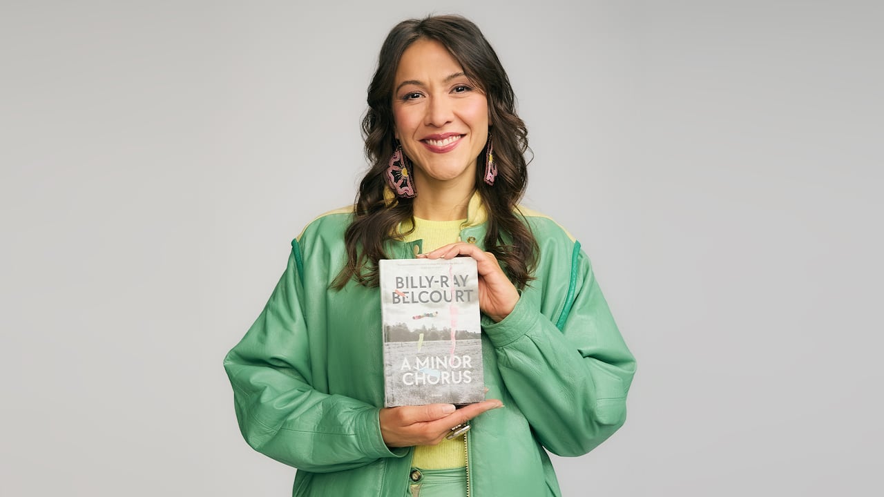 A woman with long brown and beaded earrings hair smiling hold up a book A Minor Chorus by Billy-Ray Belcourt. She has a light green jacket on and a yellow t-shirt.
