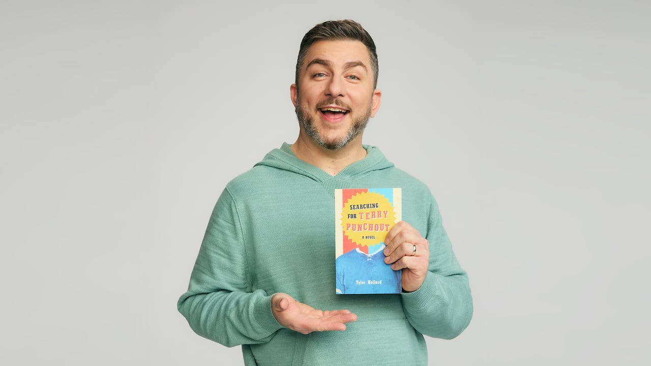 Man in green hoodie, with brown hair and greying short beard holds up the book "Searching for Terry Punchout" by Tyler Hellard.
