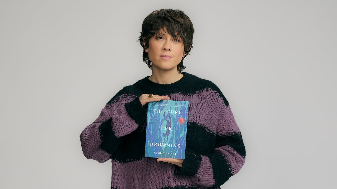 A woman with short black hair in a purple and black wool sweater holding up the book, "The Cure for Drowning" by Loghan Paylor.