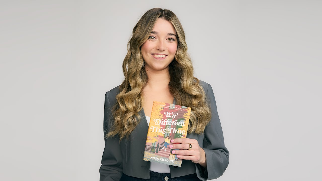 A woman with long wavy blonde hair in a white shirt and grey jacket holds up the book, It’s Different This Time by Joss Richard.