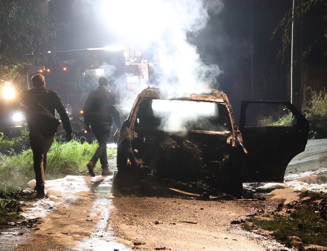 Police officers walk past a visibly smoking car.
