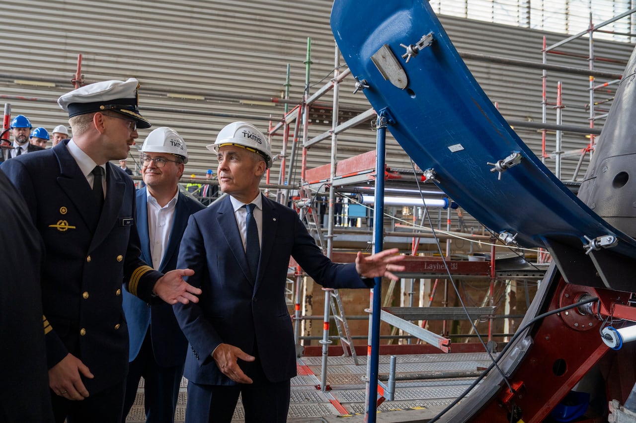 Three men stand near the propeller of a submarine, in a factory.