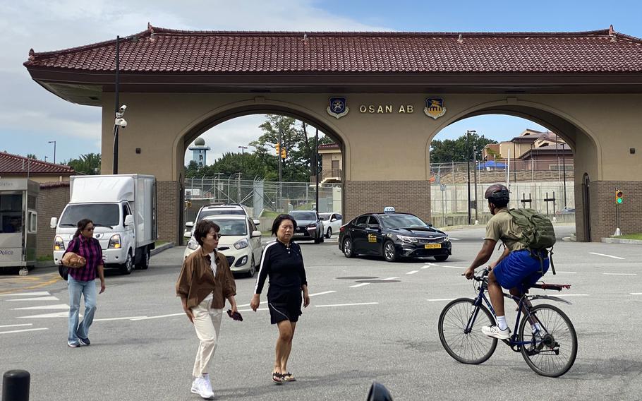 A gate at a military base, made in the style of a traditional Korean home, is seen, with cars exiting through the gate and pedestrians and a cyclist crossing the street in front.