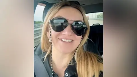 Telegram Polina Azarnykh looking straight at the camera while seated in a vehicle. She has long blonde hair and is wearing large sunglasses and a black top and is smiling.