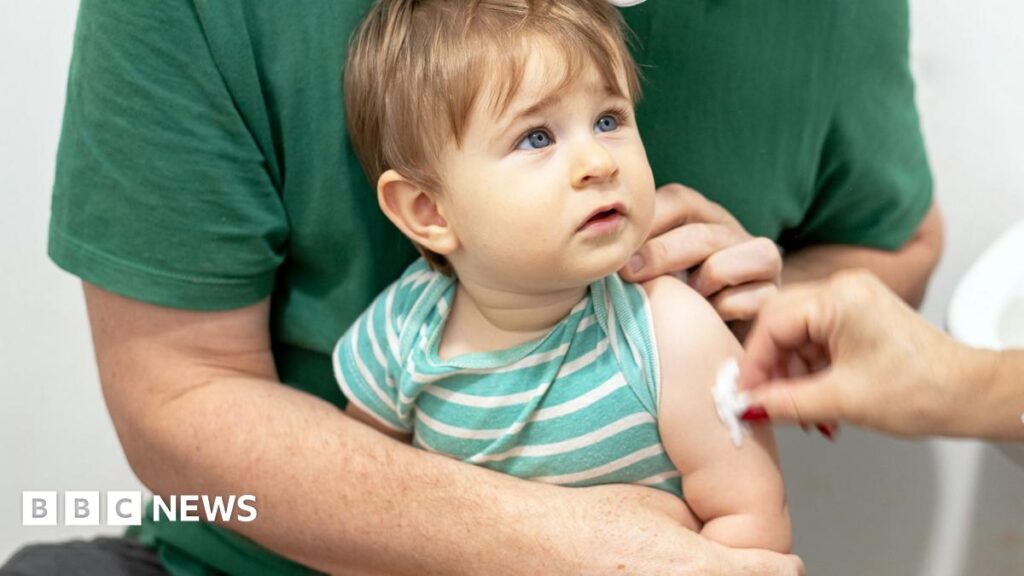 A young toddler looks up at a doctor who is holding a vaccine while rubbing a piece of cotton wool on his arm. The toddler is wearing a green and white striped T-shirt and is sitting on his father's knee, who is also wearing a green top.