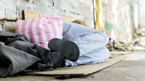 Getty Images Stock photo of a homeless person on the street. They are lying down beside a wall on a piece of cardboard with bags next to them.
