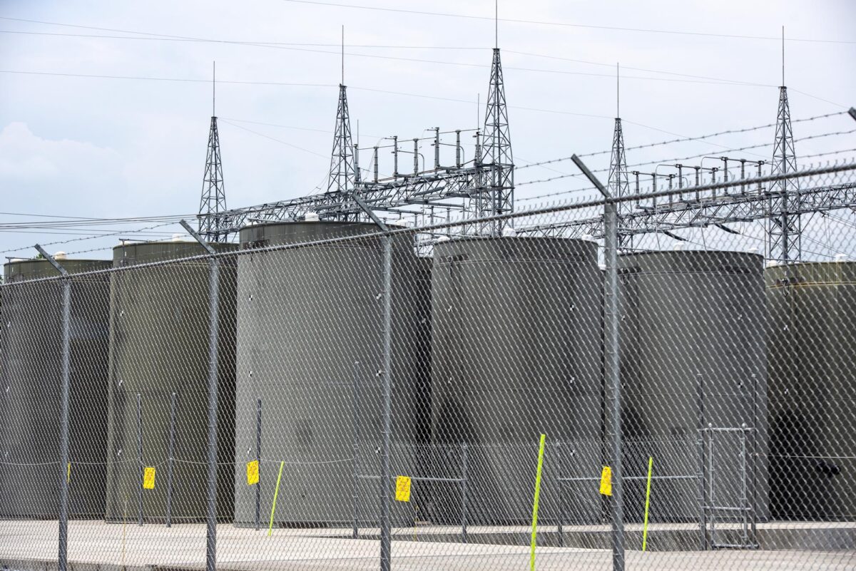 Fenced area with large cylindrical storage tanks and metal towers in the background, under a cloudy sky.