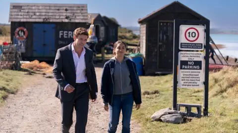 BBC/Playground Productions A man and woman walk side by side towards the camera in a publicity shot released by a production company. The man has brown hair and is wearing a suit and white shirt unbuttoned at the neck. The woman is wearing jeans, a grey hoodie and a black jacket. They are on a gravel path, with fisherman's huts behind them.
