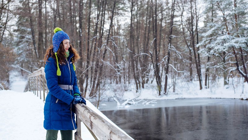 A smiling person in winter clothing standing on a snowy bridge over an icy river.