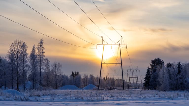 High voltage electricity wires, seen along a snowy landscape with the sun on the horizon and trees in the background.