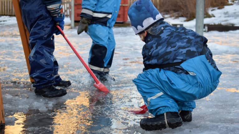 Three young children playing outdoors with red plastic shovels in the slush.