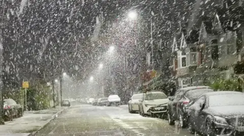 BBC Weather Watchers / Jack March A snowy street seen. Heavy snow falls from the early morning sky and is settling on a long line of parked cars that stretches back into the distance. It is also settling on the road.