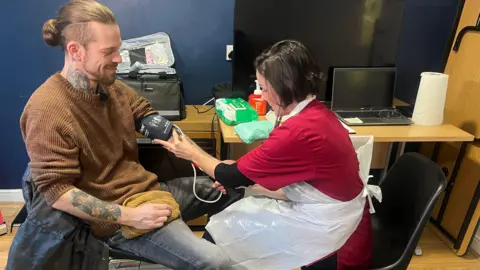 Curtis Durose having his blood pressure taken by Lisa Ewart. Curtis has long brown hair tied back, wearing a brown jumper and jeans. He has tattoos on his neck and arm. Lisa has short dark hair, wearing dark red scrubs and a disposable apron. They are sitting on black chairs beside a desk with a laptop, tissue roll, hand sanitiser and wipes on it.