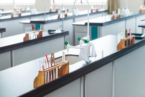 A school lab with no people but each station has equipment ready for a practical lesson including test tubes in a rack, with a holder and brush as well as a stirrer and pipette