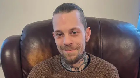 BBC Curtis Durose sitting on a brown leather armchair. Has has brown hair tied back, brown eyes, wearing a brown jumper. He has tattoos on his neck.