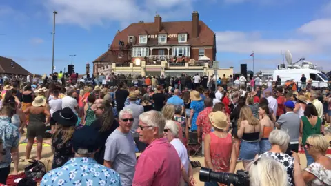 Andrew Turner/BBC A crowd of people in summer clothes standing on a beach, facing a building on the promenade. A sign across its red-tiled roof says Pier Hotel. The sky is blue.