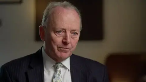 Sir Hugh Orde in a cream coloured room, he is wearing a navy pinstripe suit, a white shirt and a yellow/green floral white tie. He has short white hair.