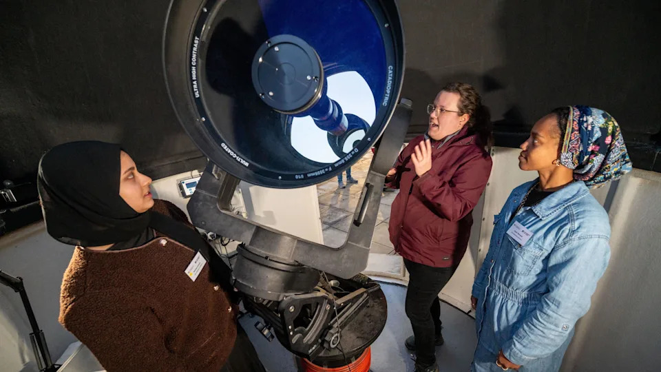Three women are stood around a telescope, which is pointing towards the left of the image.