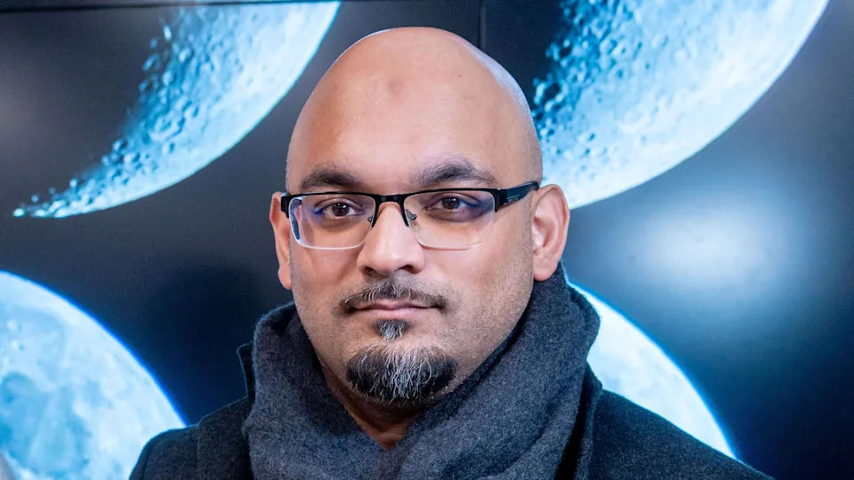 A headshot of Imad Ahmed. He is bald with a black and grey moustache and goatee beard. He is wearing glasses and a grey scarf.