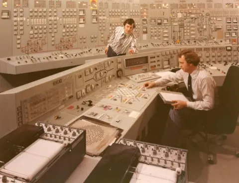 Nuclear Decommissioning Authority Inside a control room in the Hunterston B power station in the 1970s. There are two men wearing striped shirts and ties. One of them is at the control desk with a folder and a piece of paper. The other man is watching him. 