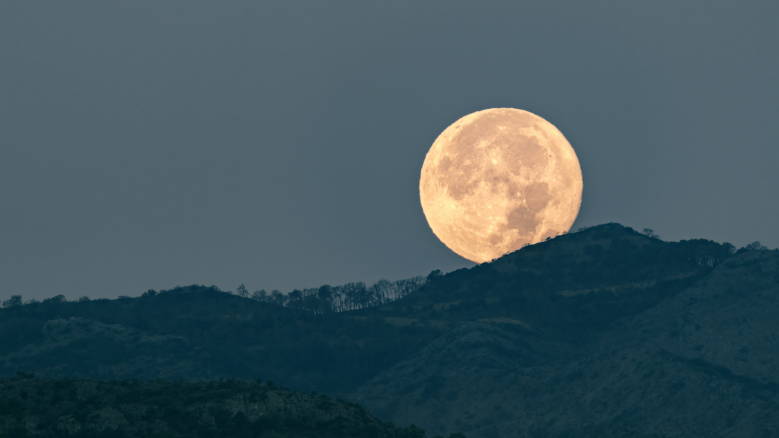 Why does the moon look larger when it’s on the horizon? This photograph freezes the exact moment of dawn when the full moon disappears behind the mountains, casting a serene and ethereal light over the landscape. Nature's quiet spectacle unfolds as the moon bids its silent farewell against the backdrop of the waking world.