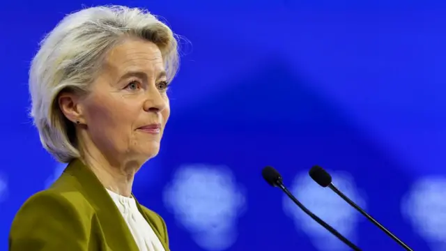 Ursula von der Leyen, a white lady with blonde hair, wearing a green jacket and white shirt standing behind microphones.