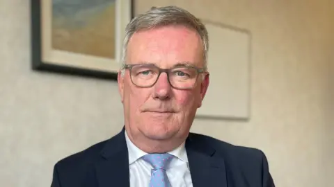 PA Media A close-up image of Mike Nesbitt. He is an older man with short, grey hair. He is wearing a pair of grey frame glasses, and is wearing a dark suit jacket, a white hsirt and a pale blue tie with pink spots. He is looking at the camera with a neutral expression on his face as he sits in a cream coloured room.