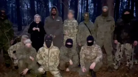 Telegram Azarnykh in a dark coat standing next to nine people in military clothing, with dense forest in the background. Next to her is Habib, who is wearing a khaki-coloured hood but his face is visible. The other people's faces are covered with black balaclavas or scarves, except for one whose face has been blurred. 