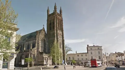 Google A gothic style church with a square tower at one end, stands on a town street with green-leafed trees in front of it. Beside the church are houses on both sides and a bus is running along a town street away from us.